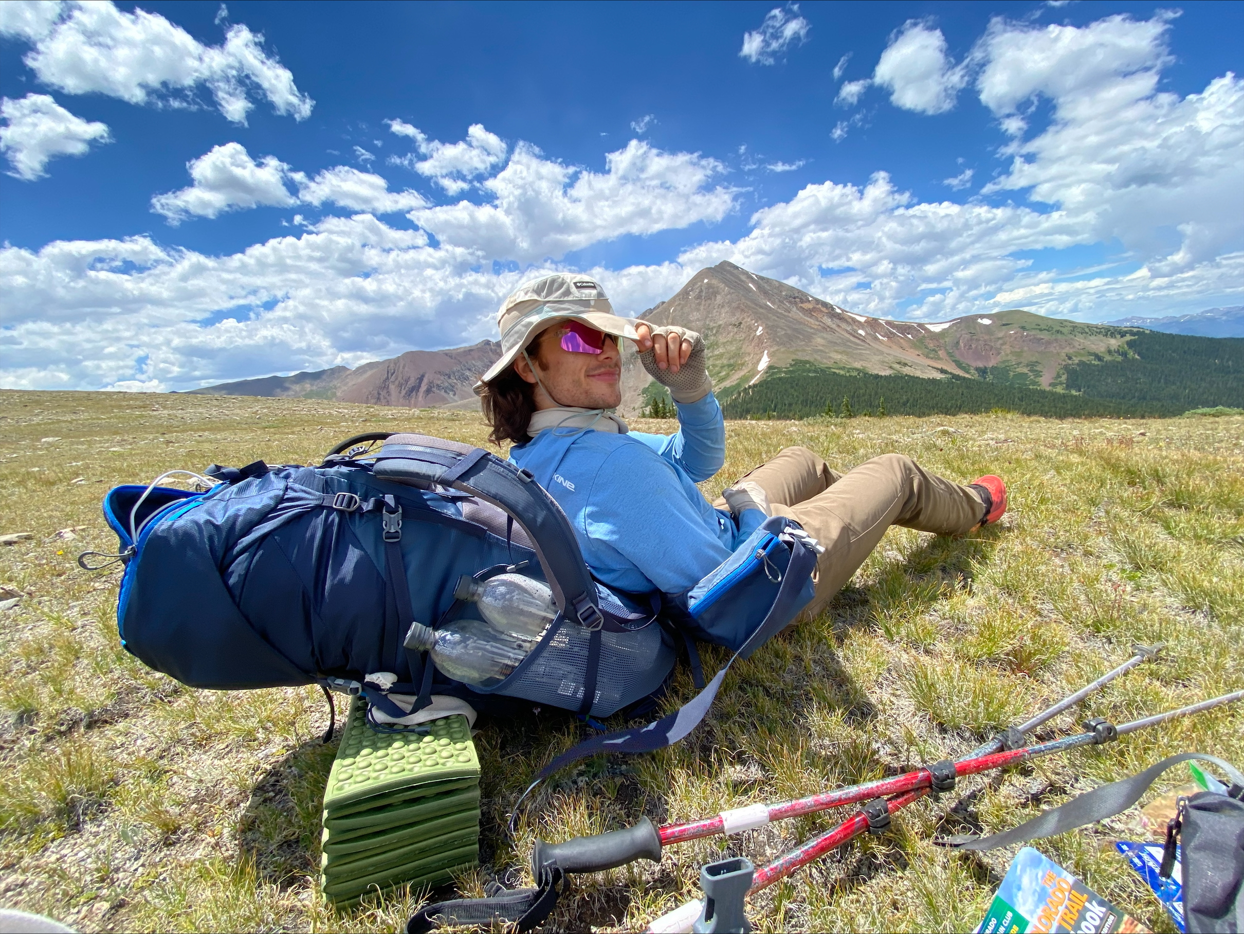 Henry with hat and sunglasses leaning against backpack in field in front of mountain.
