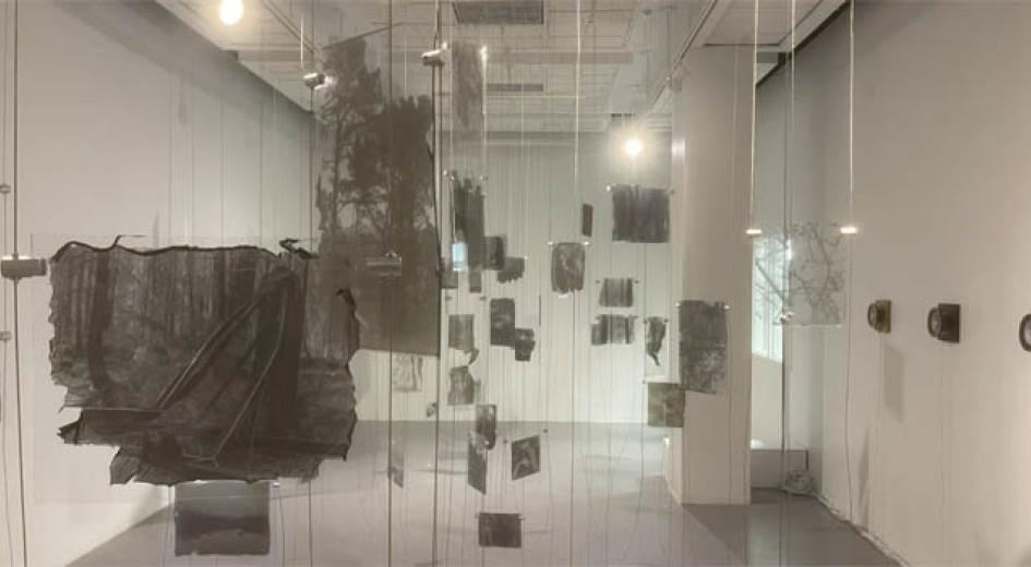 Gallery with black and white photos printed on glass suspended from the ceiling and speakers mounted on the wall. 