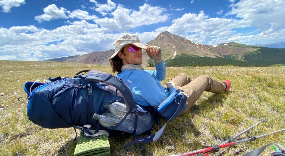 Henry with hat and sunglasses sitting in field leaning against backpack with mountain in background.