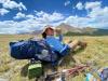 Henry with hat and sunglasses sitting in field leaning against backpack with mountain in background.